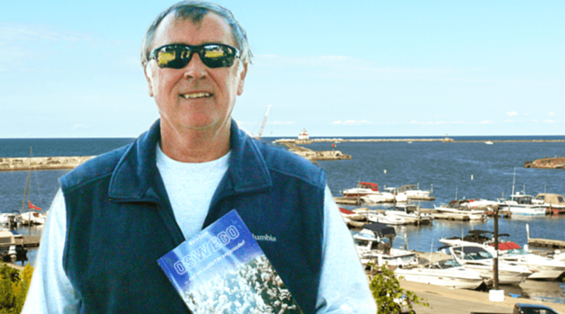 Mike McCrobie, with the Oswego lighthouse in the background, holds his second book — ‘We’re from Oswego ... and we couldn’t be any prouder!’ — which was published in November.